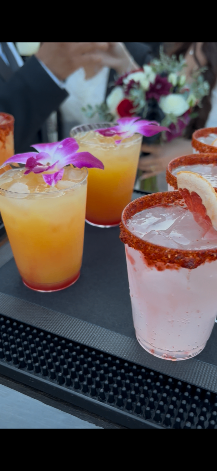 Colorful orchid-garnished cocktails with chamoy rims lined up on the bar mat at a wedding