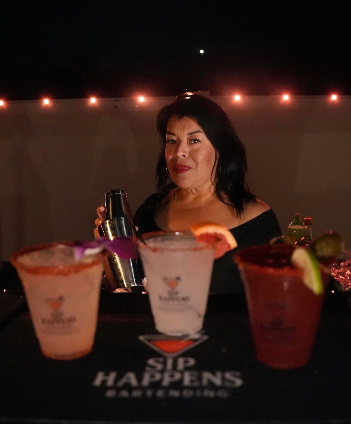Sip Happens bartender with a shaker behind three chamoy-rimmed cocktails at a night event
